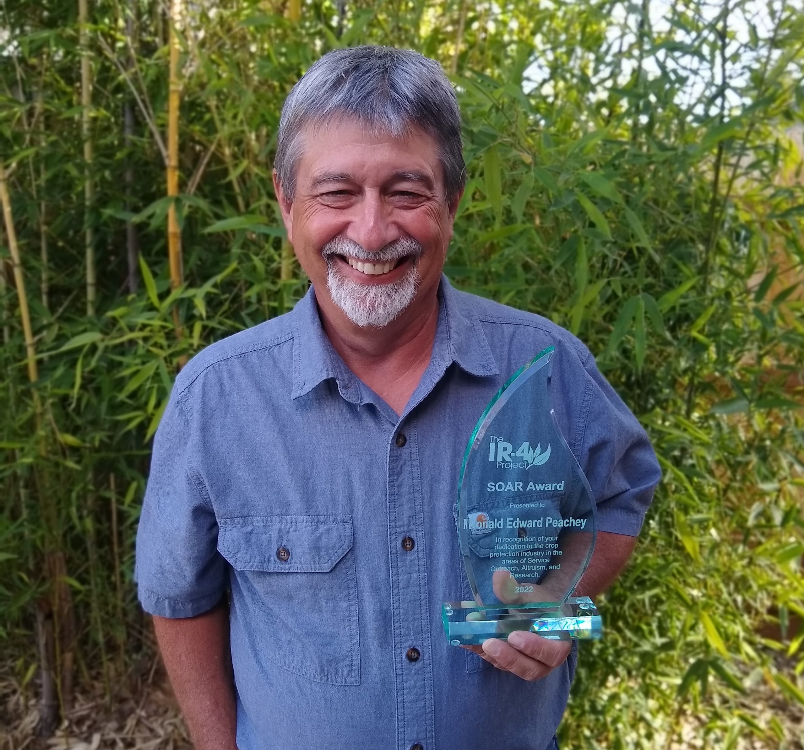 Man standing outside in front of large plants, wearing a blue button-up, short sleeve shirt and holding an award that says "IR-4 Project SOAR Award Ronald Edward Peachey"