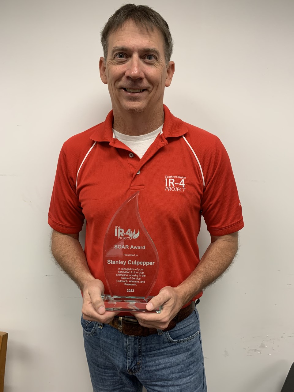 Man wearing a read shirt holding an award that says "IR-4 Project SOAR Award. Stanley Culpepper"