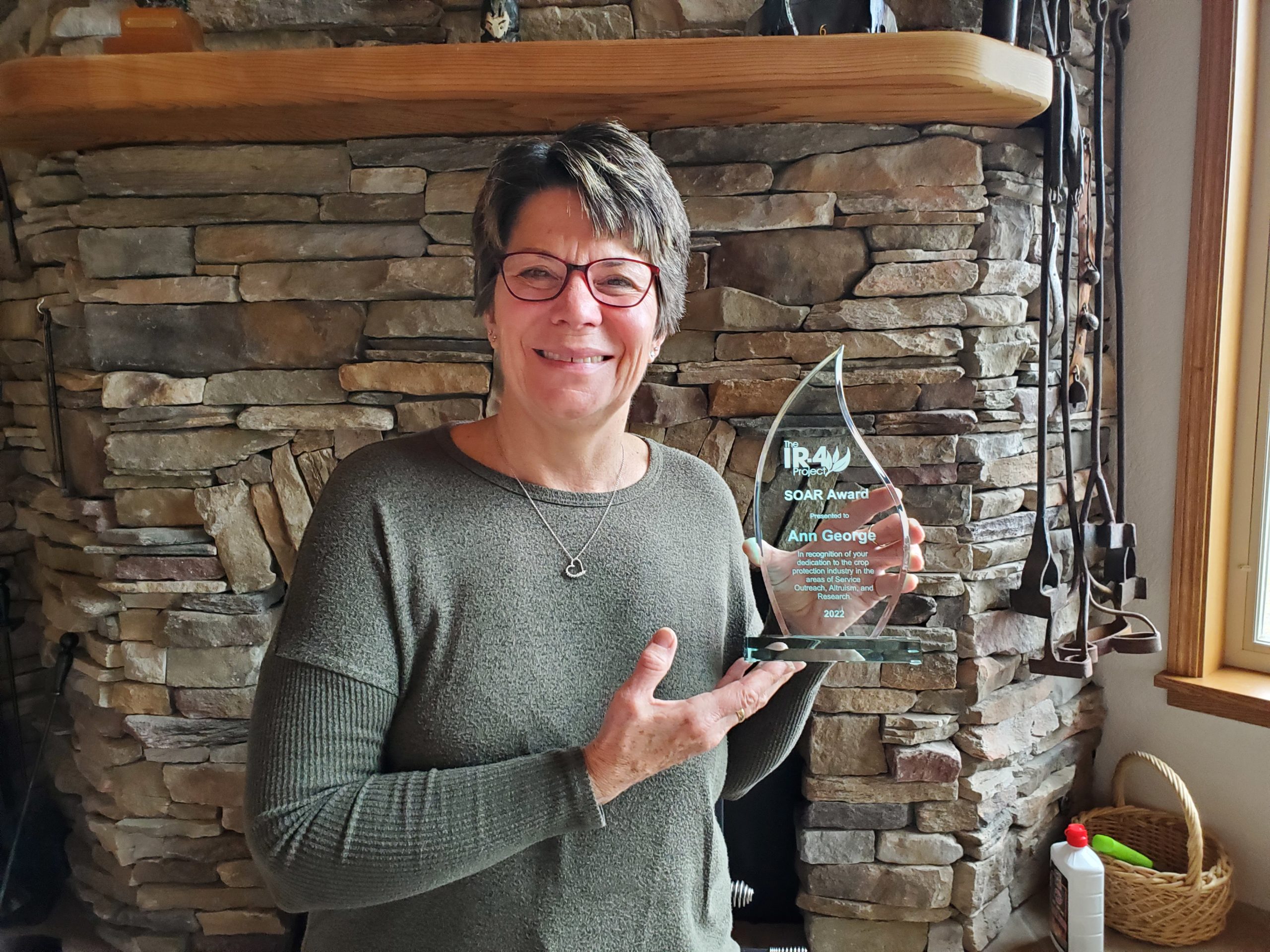 Woman with short hair and glasses standing in front of a fireplace and holding an award that says "IR-4 Project SOAR Award presented to Ann George"